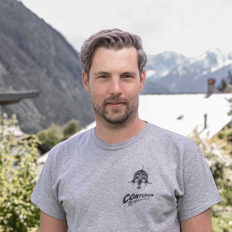 Mann mit kurzen Haaren und Bart im grauen T-Shirt steht draußen vor den Bergen im Hintergrund.