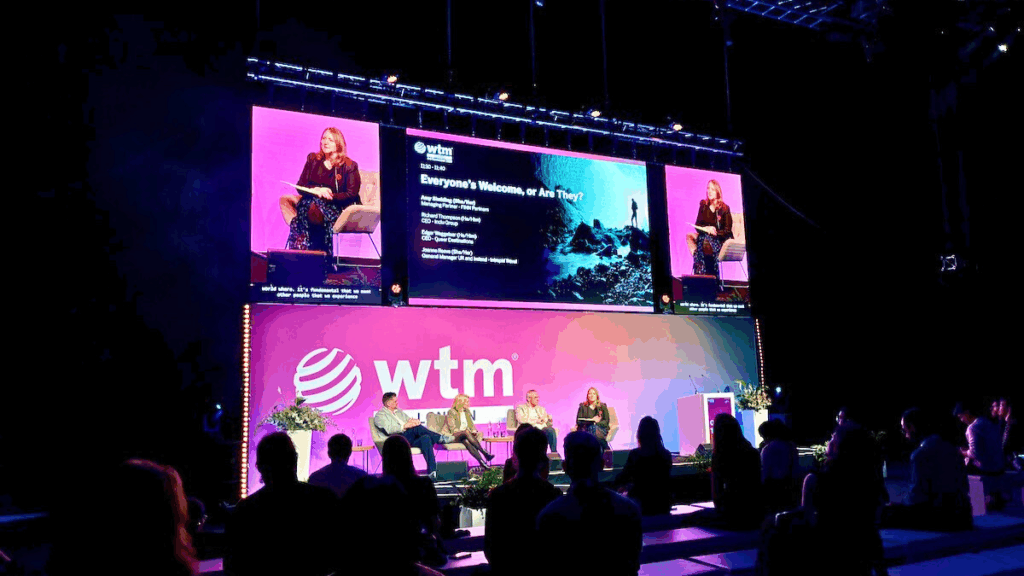 Podiumsdiskussion auf der Bühne der WTM-Veranstaltung mit Zuschauern und Großbildschirm im Hintergrund.
