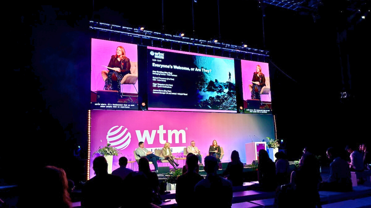 Podiumsdiskussion auf der Bühne der WTM-Veranstaltung mit Zuschauern und Großbildschirm im Hintergrund.